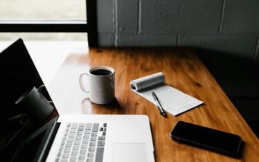 Wooden desk featuring a laptop, a cup of coffee, a notepad with a pen, and a smartphone, symbolising Faith and Work Balance.