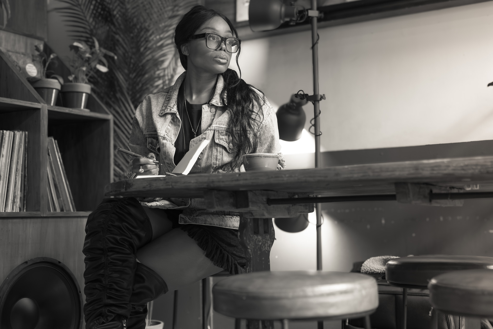 Black-and-white portrait of a woman seated at a table, writing in a notebook, looking away thoughtfully in a café setting.